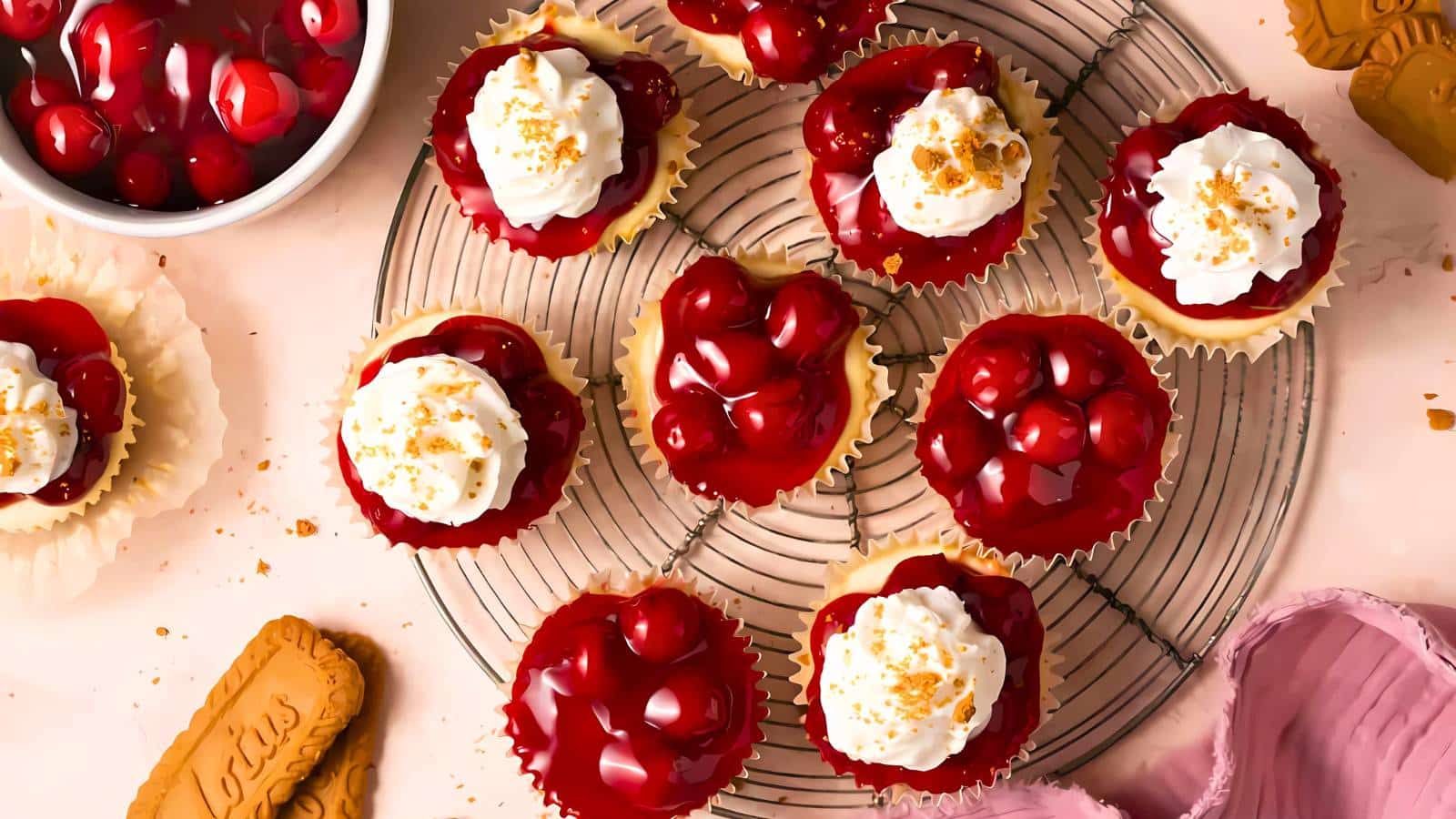 A rack of cherry-topped cupcakes with whipped cream and crumb decoration. Additional cupcakes, a bowl of cherries, and scattered crumbs are also visible around the rack.