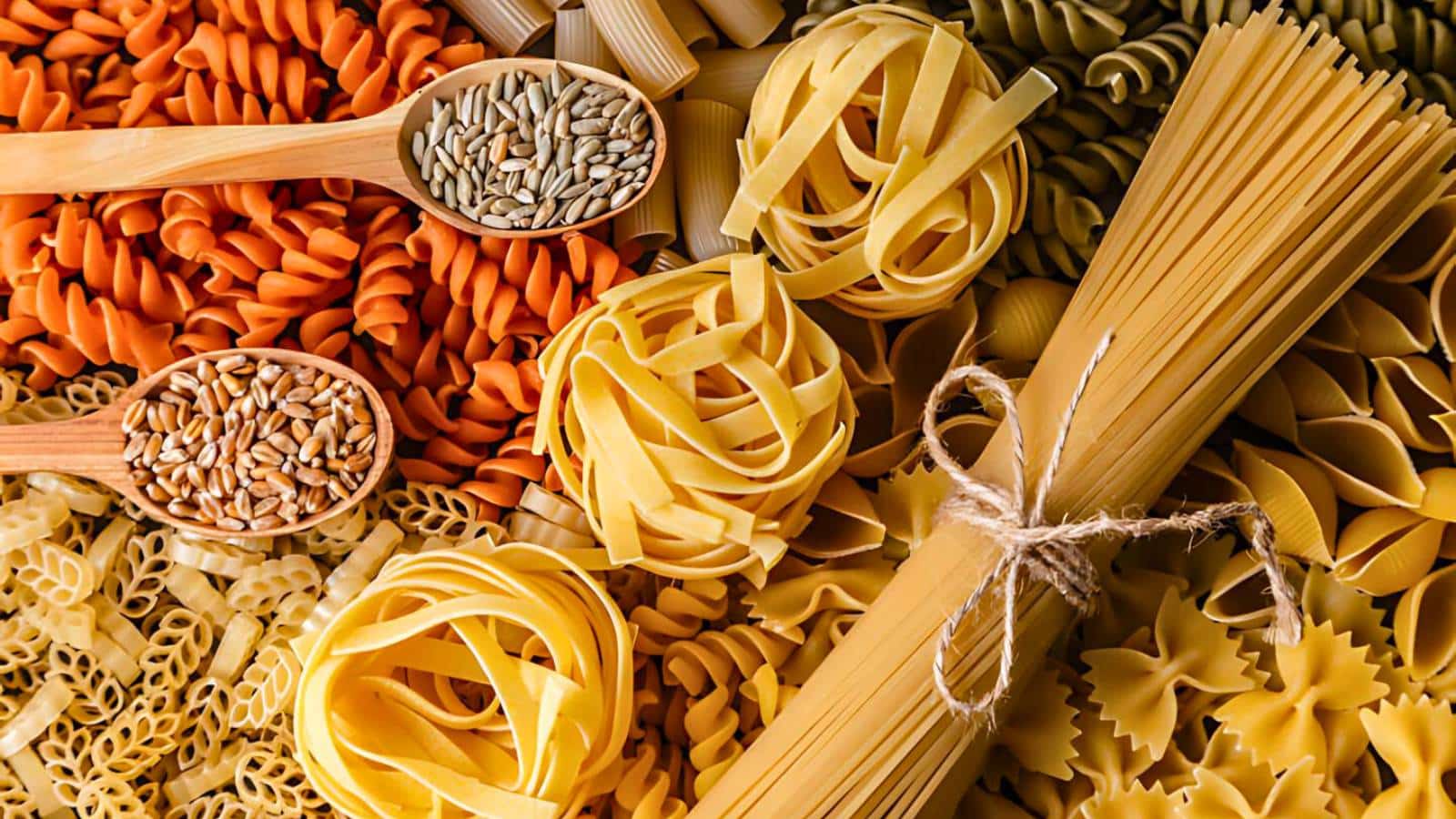 An assortment of uncooked pasta varieties and seeds arranged with wooden spoons, including fusilli, tagliatelle, penne, and bow-tie pasta tied with string.