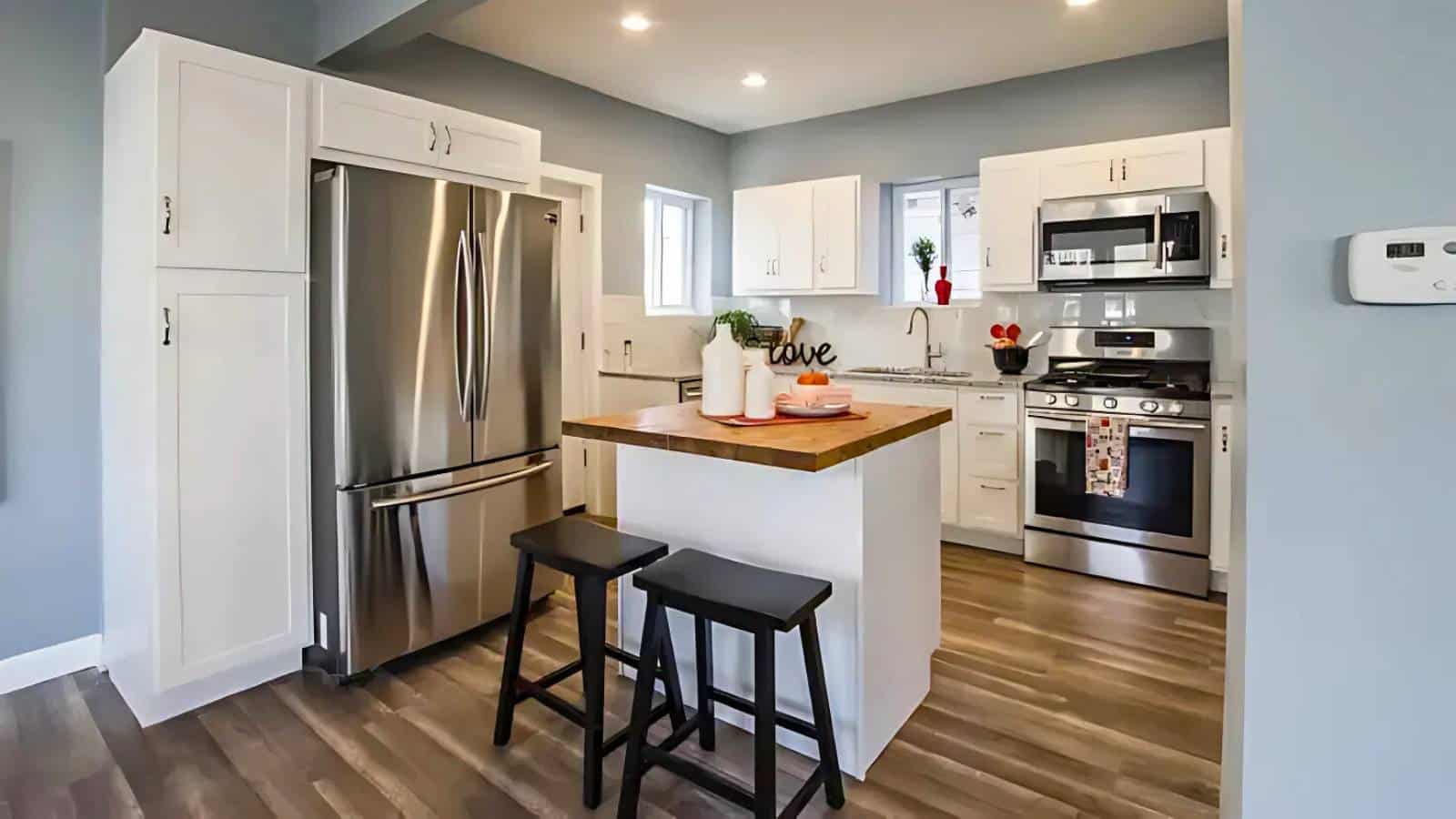 Modern kitchen with white cabinets, stainless steel appliances, and an island with two stools. Light wood flooring and recessed lighting complete the space.