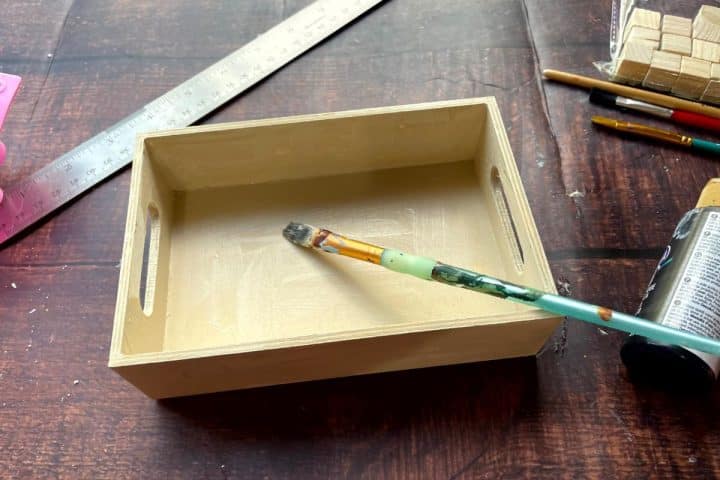 A wooden tray being painted with a brush, surrounded by a ruler, colored pencils, small wooden blocks, and a tube of black paint on a wooden table.