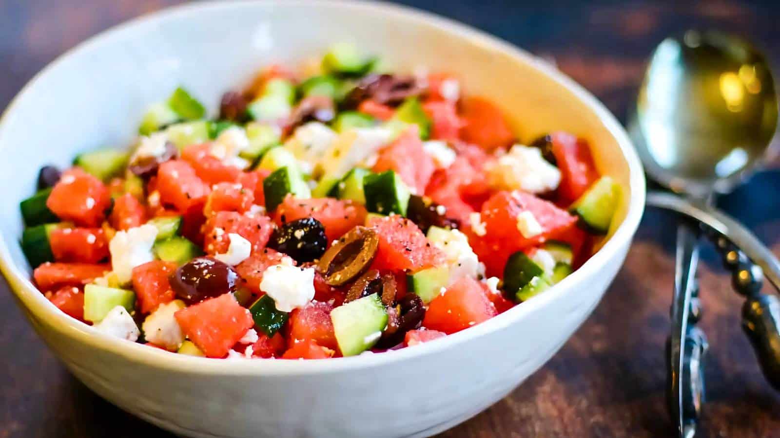 A bowl of cubed watermelon, cucumber, feta cheese, and olives is on a wooden surface, with a spoon and fork beside it.