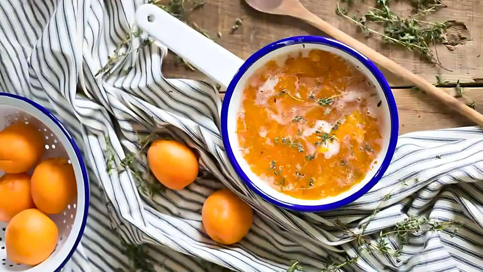 A white saucepan filled with an orange mixture, garnished with herbs, sits on a striped cloth napkin next to whole apricots and a wooden spoon.
