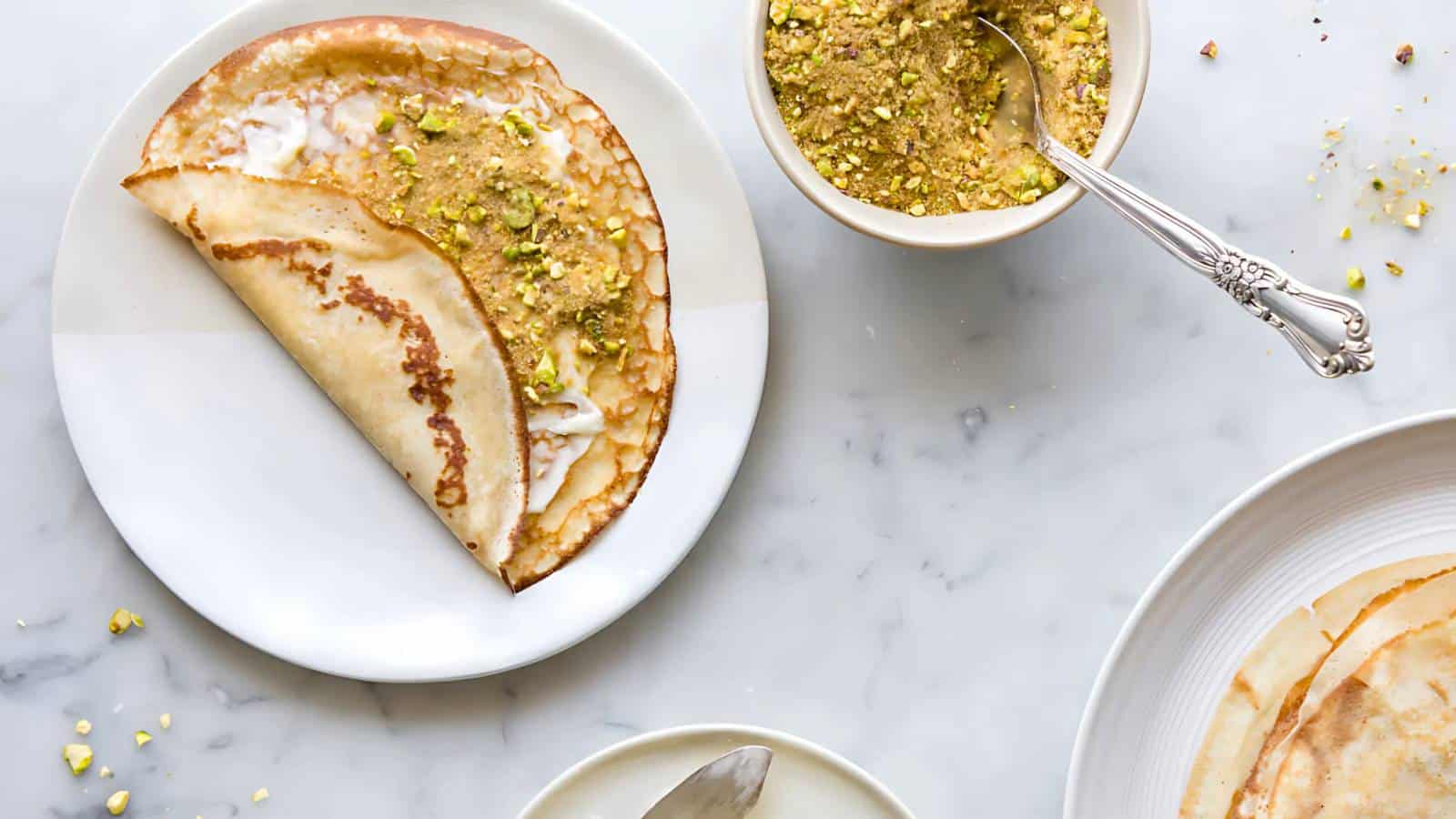 A crepe folded on a plate is topped with crushed pistachios. A bowl of pistachios and other crepes are on a marble surface.