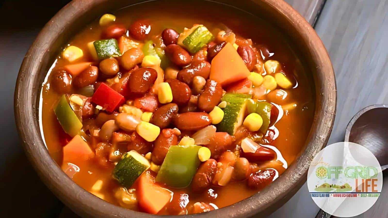 A bowl of hearty vegetable and bean soup, simmering with corn, bell peppers, carrots, and beans—perfected as a woodstove recipe—with an "Off Grid Life" logo in the corner.