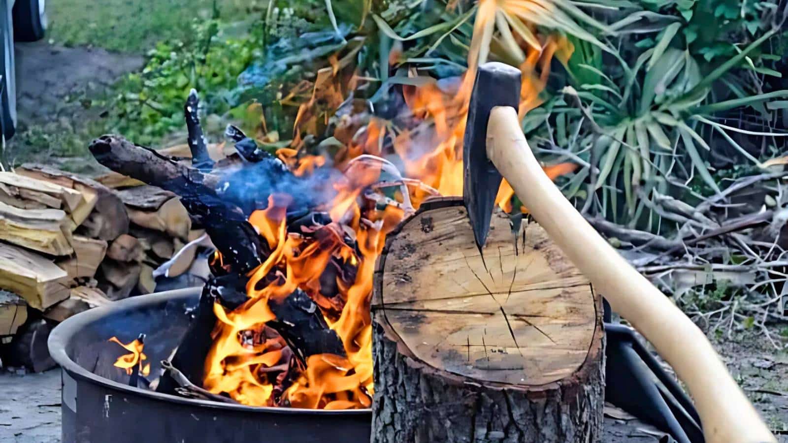 A log burning in a fire pit with an axe embedded in a tree stump nearby. Stacked firewood and greenery are in the background.