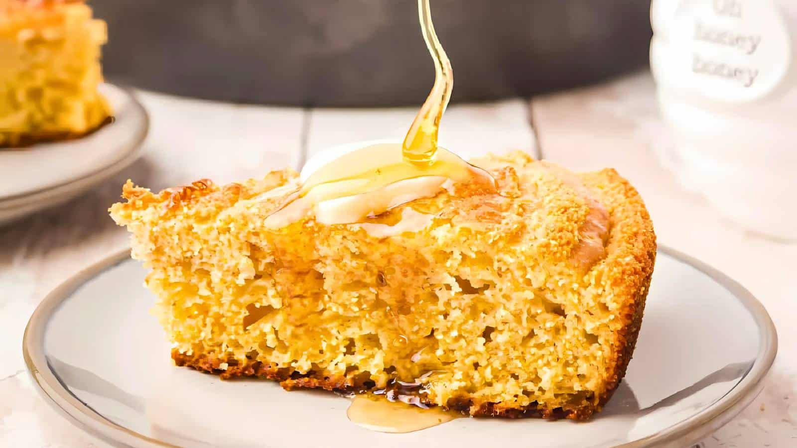 A slice of cornbread on a plate topped with a dollop of whipped butter and a drizzle of honey.