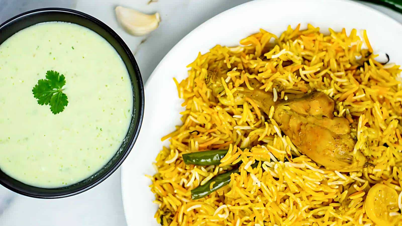 A plate of biryani with chicken and vegetables next to a bowl of green yogurt sauce garnished with cilantro.
