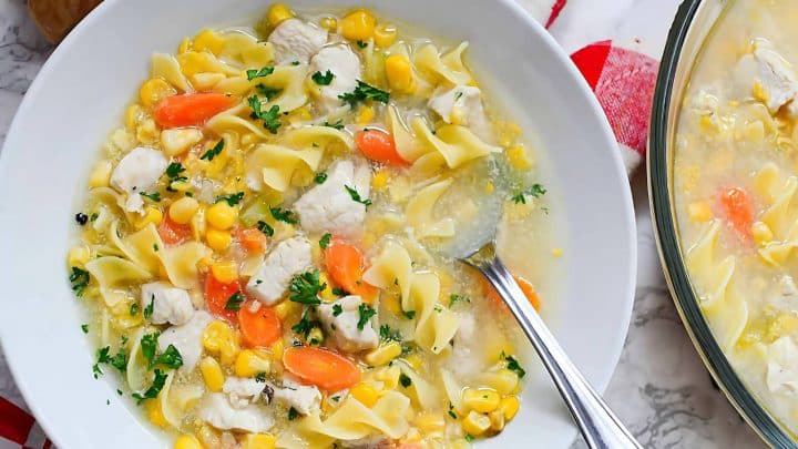 A bowl of chicken noodle soup with corn, carrots, and parsley, served with a spoon.