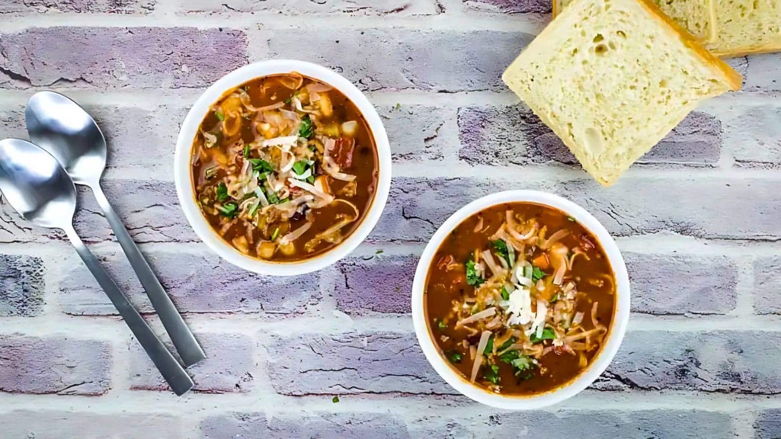 Two bowls of soup topped with cheese and herbs are placed on a textured surface, accompanied by two spoons and slices of bread.