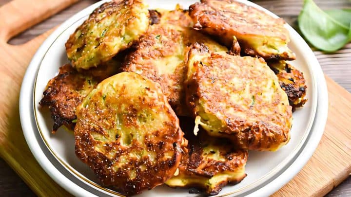 Plate of golden-brown vegetable fritters stacked on a white dish, resting on a wooden surface, with a leaf garnish slightly visible in the background.