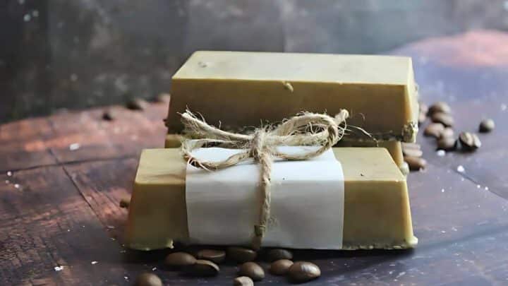 Two rectangular bars of soap, one wrapped with twine and paper, placed on a wooden surface with coffee beans scattered around.