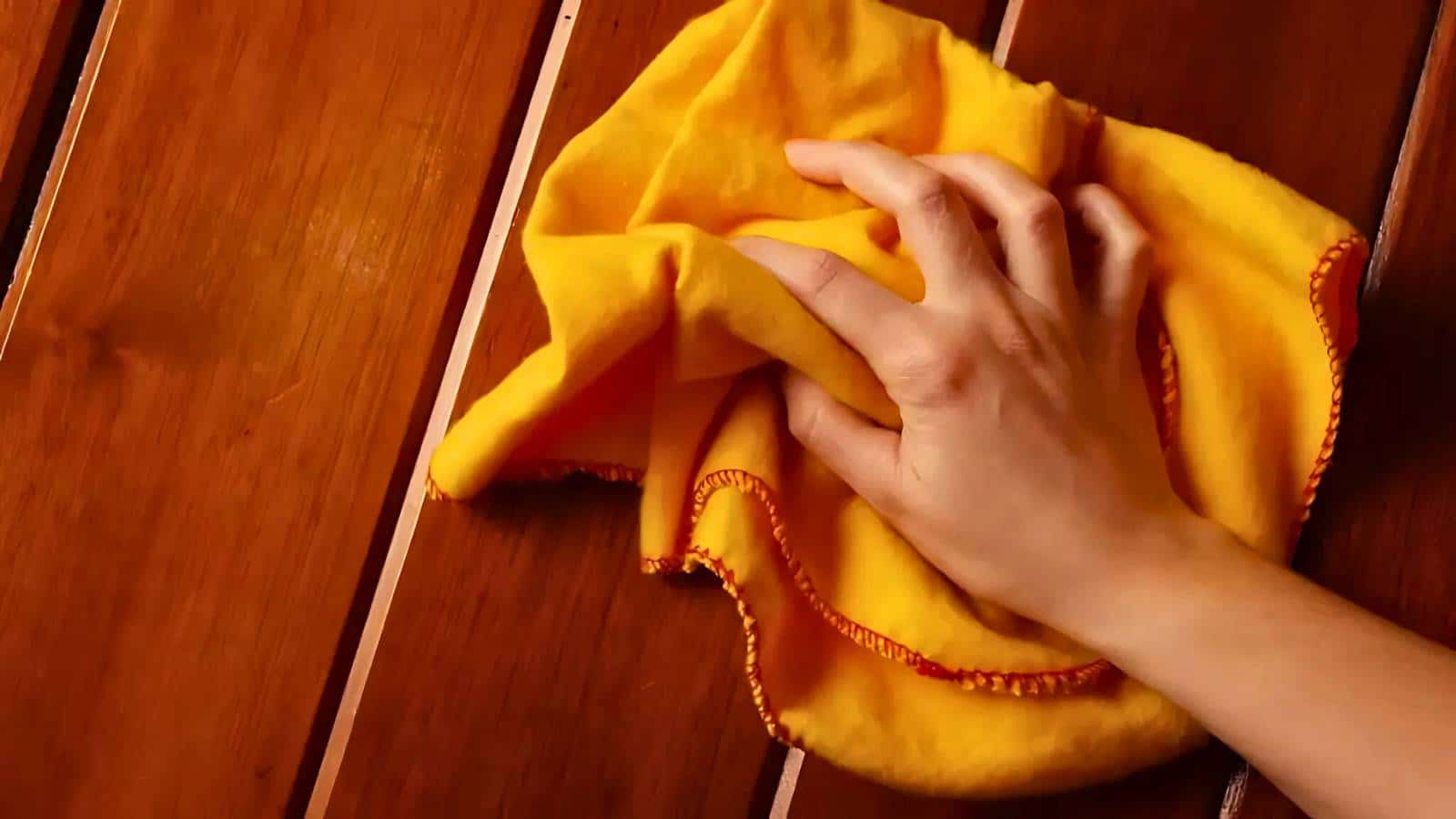 Hand wiping a wooden surface with a yellow cloth.