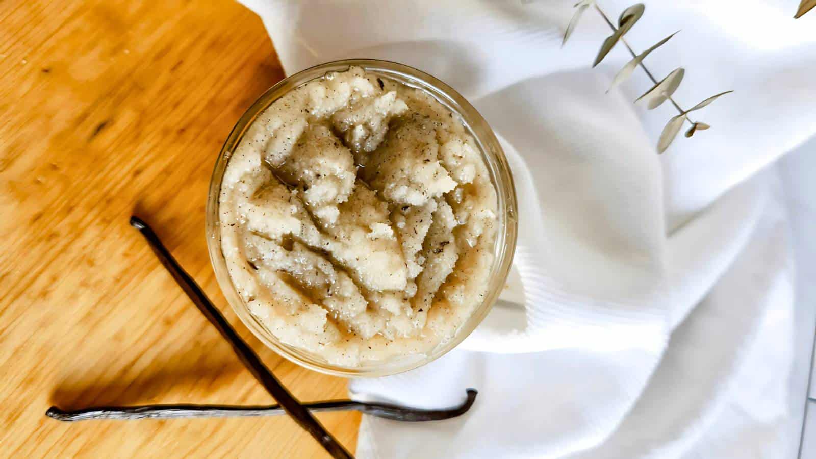 A jar filled with a creamy mixture is on a wooden surface. A vanilla bean pod and a white cloth are nearby.