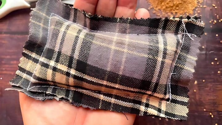 A hand holds a small handmade pouch made of black and beige plaid fabric, with uneven edges and visible white stitching, over a wooden surface with scattered grains.