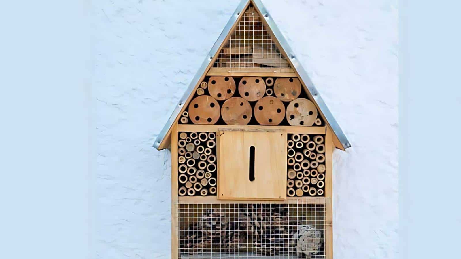 A wooden insect hotel with various cylindrical compartments and drilled holes, mounted on a white wall.
