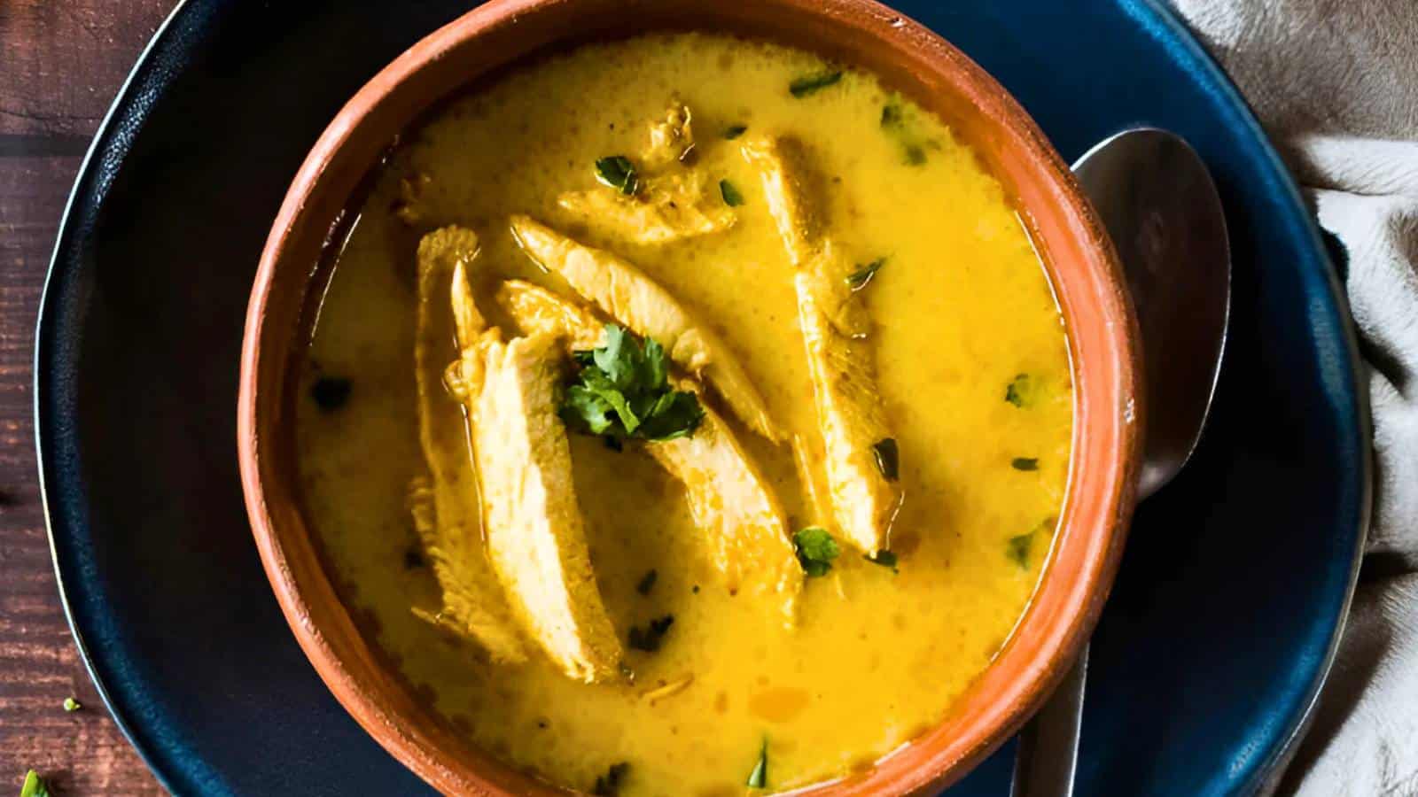 A bowl of yellow curry with sliced chicken and cilantro, served in a brown dish on a blue plate, with a spoon beside it.