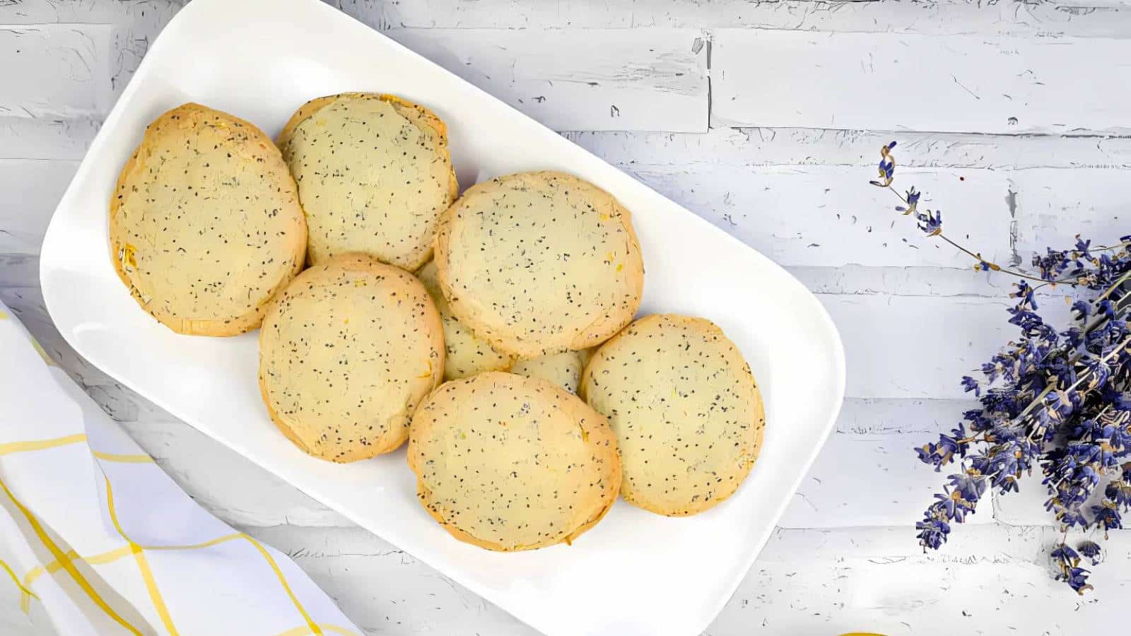 A white rectangular plate with seven poppy seed cookies on a gray wooden surface, next to a yellow and white cloth and dried purple flowers.