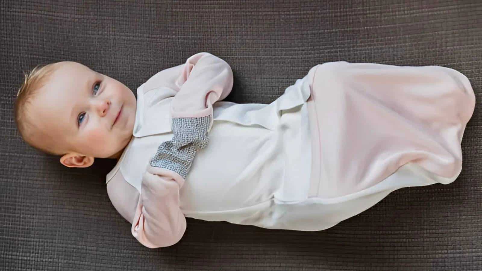 A baby in a white and pink outfit with gray mittens is lying on a dark textured surface, smiling.