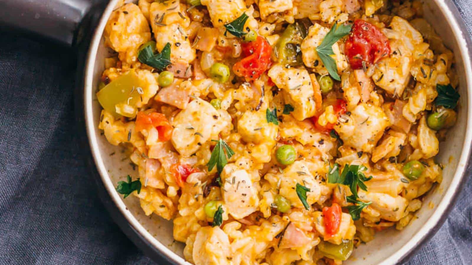 A bowl of mixed risotto with diced chicken, green peas, tomatoes, bell peppers, and parsley garnish on a dark cloth background.