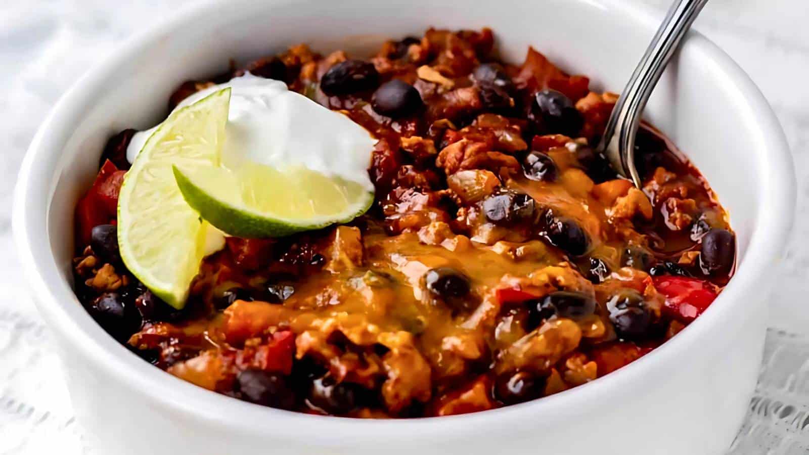 A bowl of chili with black beans, melted cheese, and a spoon, garnished with a lime wedge and a dollop of sour cream.