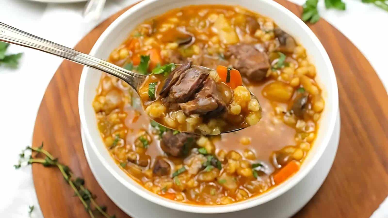 Bowl of beef barley soup with carrots, mushrooms, and herbs. A spoon holds a portion above the bowl.
