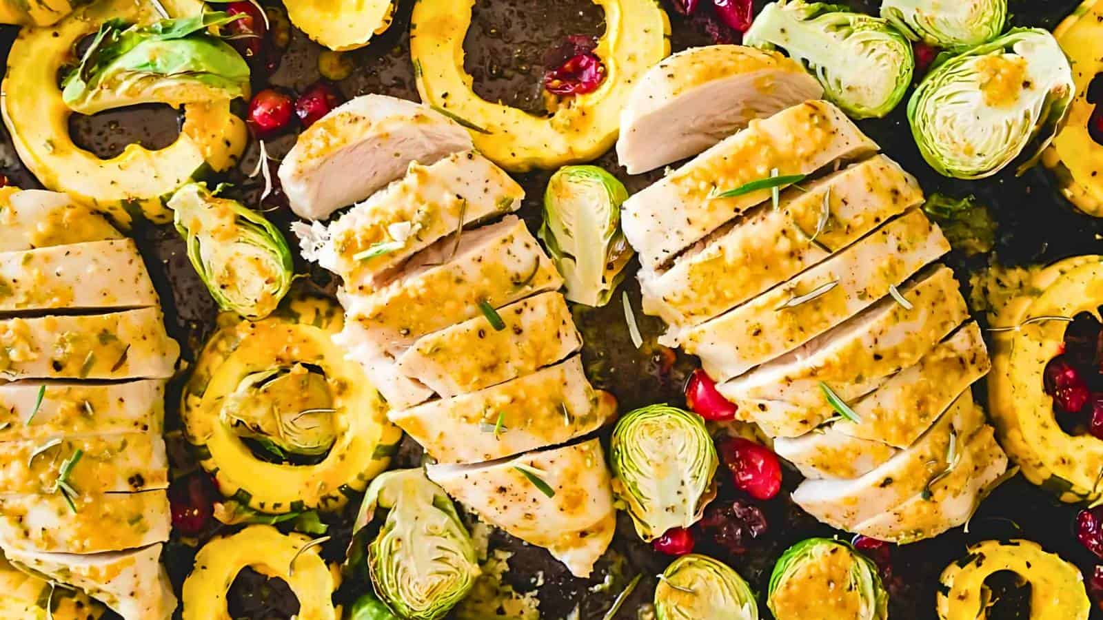 Sliced chicken breasts with rosemary on a baking sheet, surrounded by roasted Brussels sprouts, squash rings, and scattered pomegranate seeds.