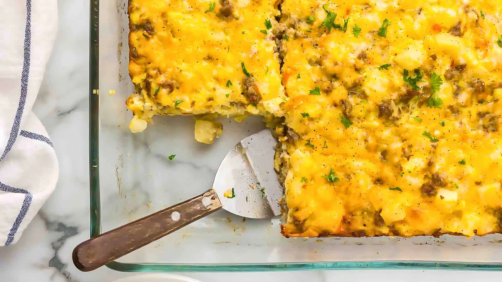 A baked casserole with a slice removed, revealing cheese, ground meat, and herbs. A metal spatula rests in the empty section of a glass dish on a marble surface.