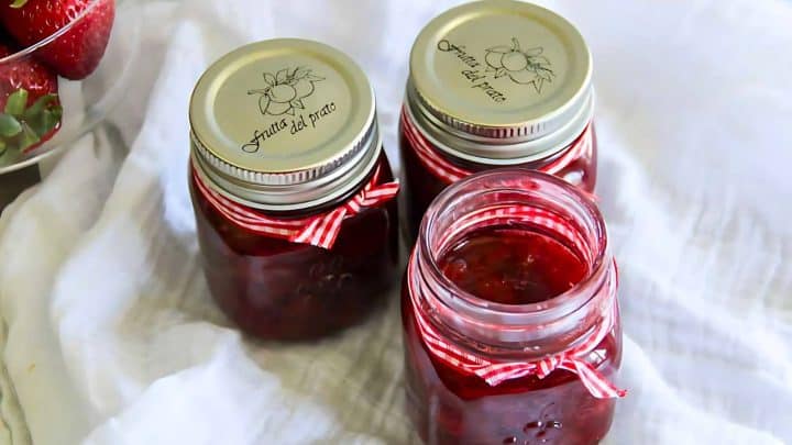 Three glass jars filled with red jam, each with a silver lid and a red-and-white striped ribbon, are placed on a white cloth.