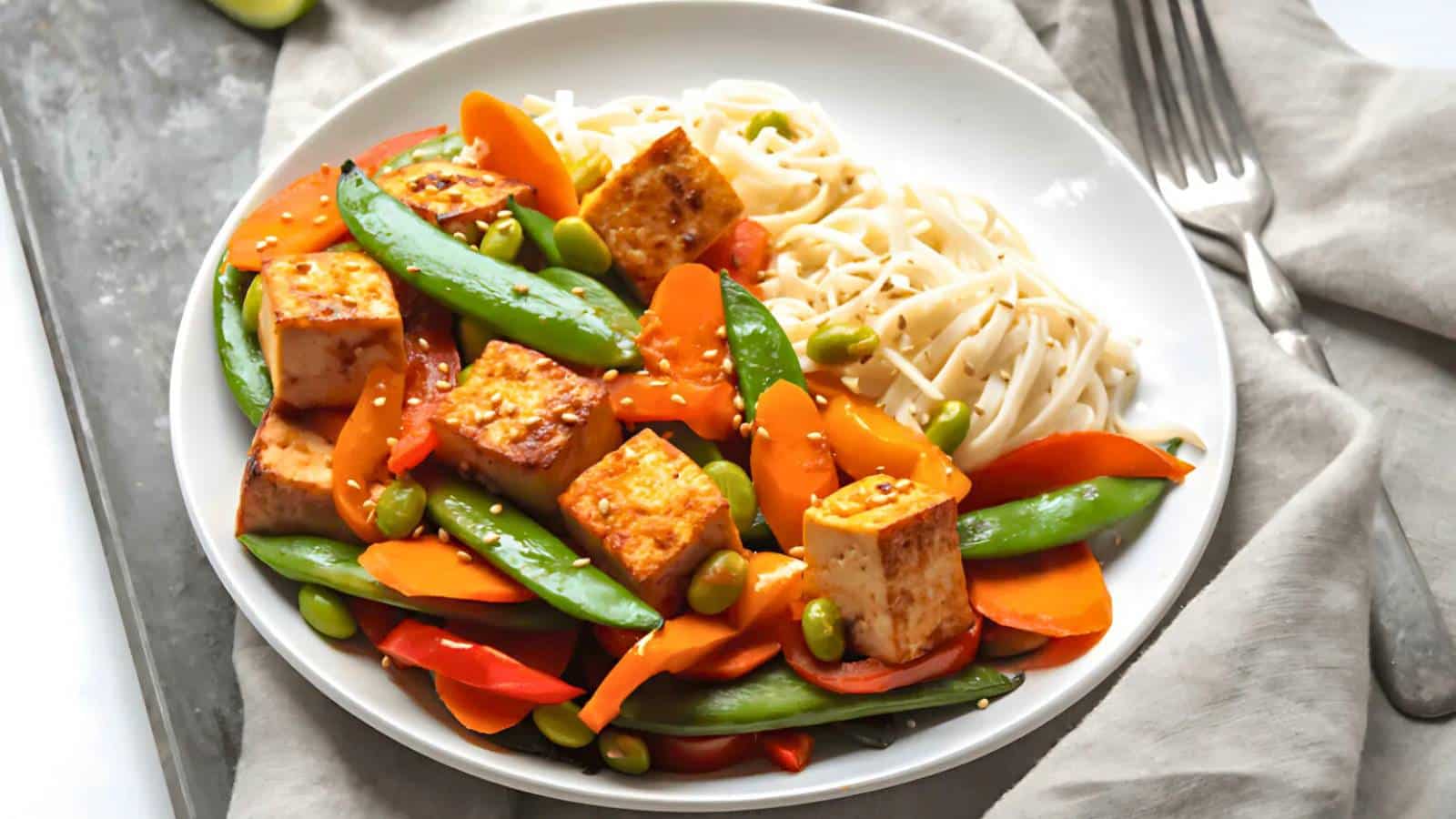 A plate with tofu cubes, bell peppers, snap peas, edamame, and udon noodles, garnished with sesame seeds. A fork and napkin are beside the plate.