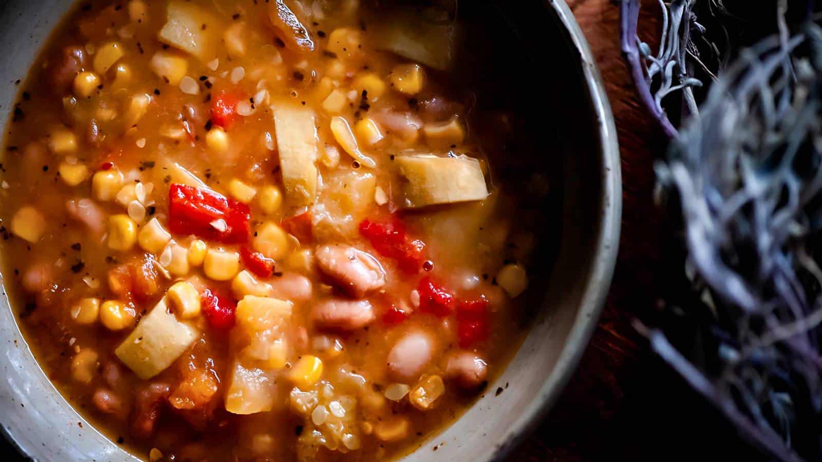 A bowl of hearty stew with beans, corn, red peppers, and chunks of potato in a thick broth.