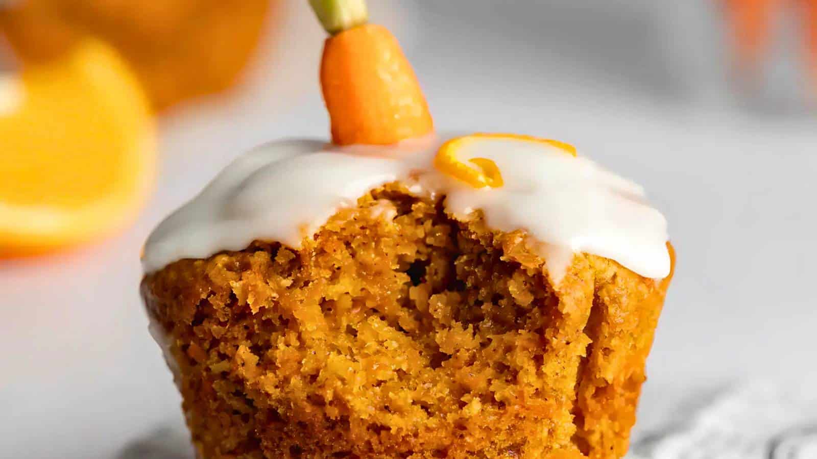 Close-up of a carrot cake muffin with white icing, topped with a small carrot-shaped piece. A bite is taken from the muffin, and an orange slice is blurred in the background.