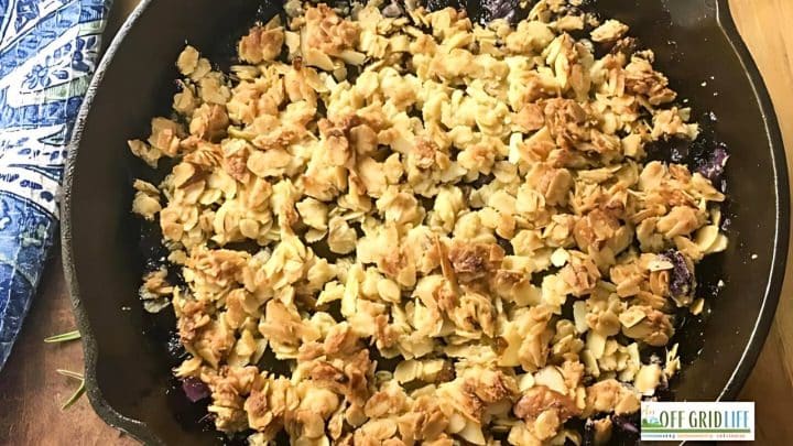 A close-up of a skillet with a freshly baked dessert topped with a golden, crumbly mixture beside a folded blue patterned cloth. The Off Grid Life logo is visible in the bottom right corner.