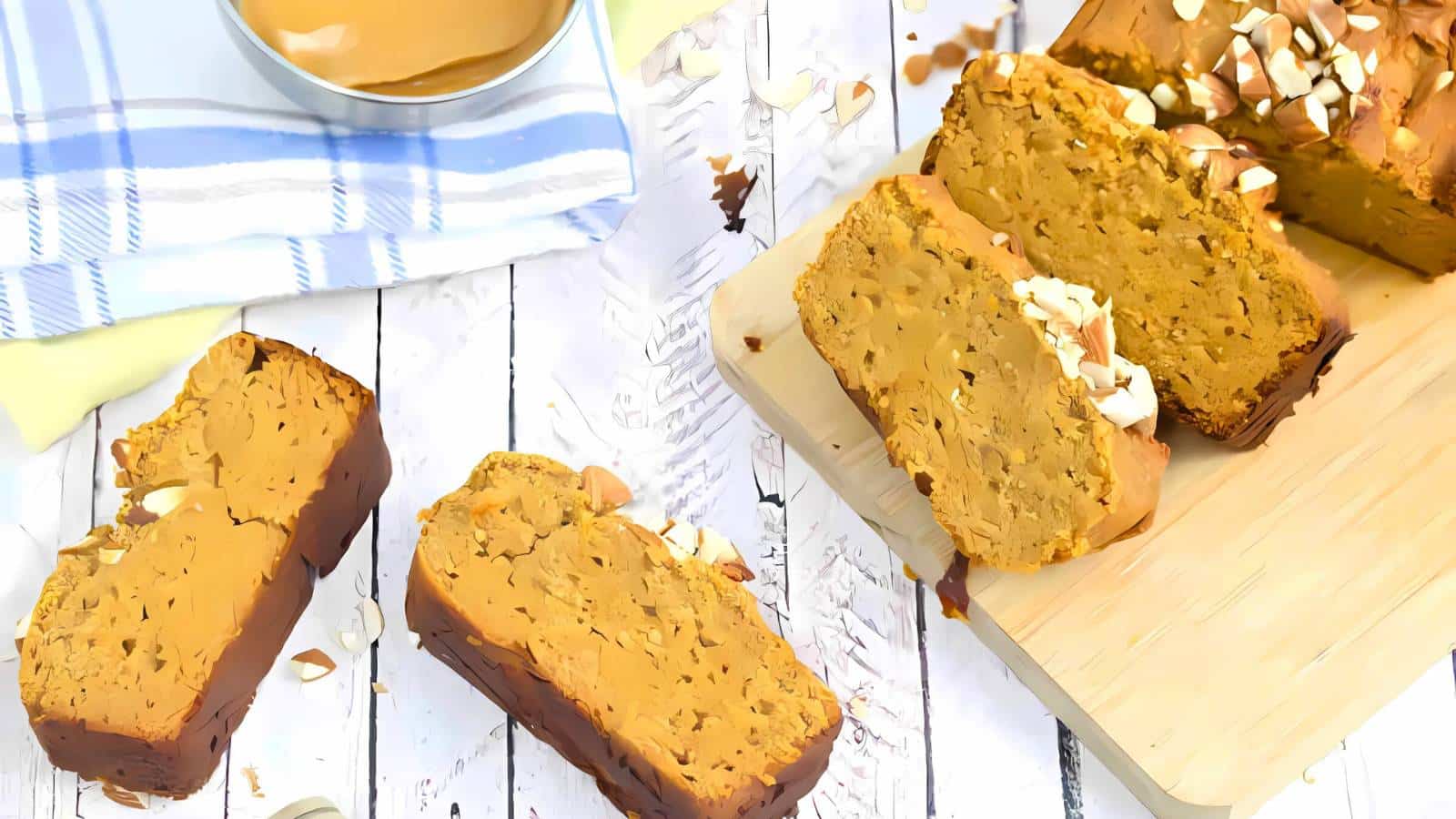Sliced loaf of nut bread on a wooden board, with additional slices on a white rustic surface. A blue-striped cloth and a bowl of spread are nearby.