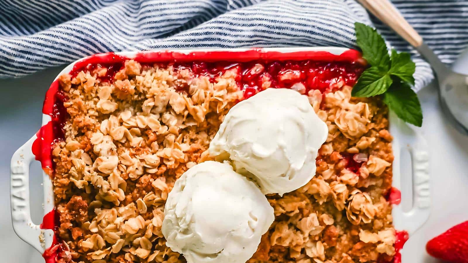 A tray of berry crumble topped with two scoops of vanilla ice cream, garnished with fresh mint. A striped cloth and a wooden spoon are in the background.