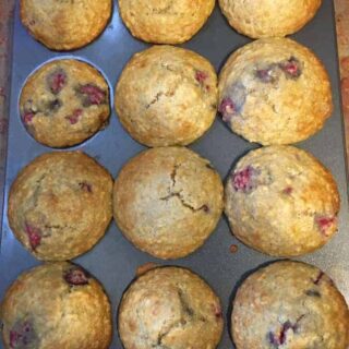 A muffin tin holds twelve golden-brown muffins, some with visible berry pieces-one compartment is empty, hinting at a popular recipe for raspberry lemon almond muffins.