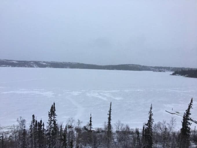 A frozen lake is surrounded by snow-covered trees and distant hills under a gray, overcast sky—an off grid mama’s winter haven.