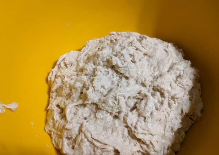 A close-up of a rough, partially mixed blueberry bread dough in a yellow mixing bowl.