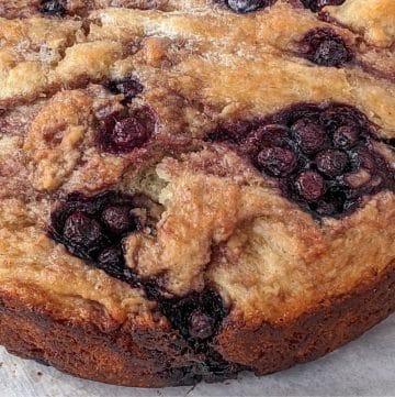 A close-up of a round baked cake with a golden crust and visible clusters of blueberries throughout.
