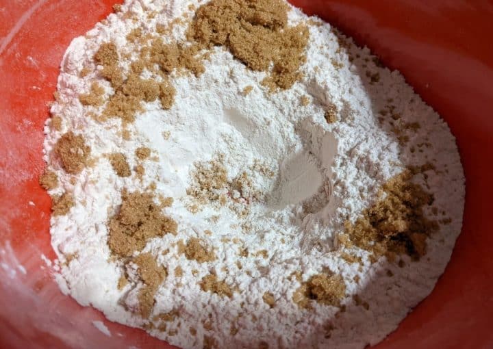 A red mixing bowl containing flour and scattered brown sugar.