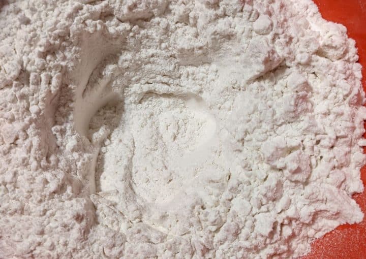 A close-up of white flour with a well in the center, in a red mixing bowl.