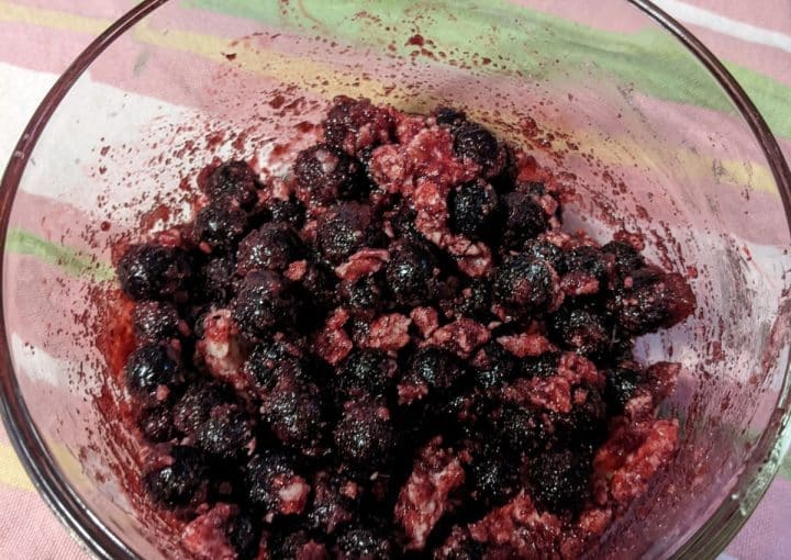 A glass bowl containing a mixture of dark berries and what appears to be sugar, set on a pink and green striped tablecloth.