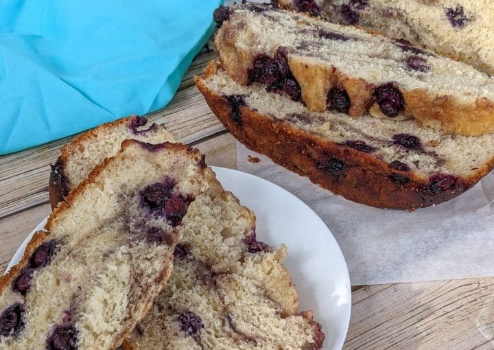 Sliced loaf of bread with purple berries baked inside, served on a white plate next to a blue cloth on a wooden surface.