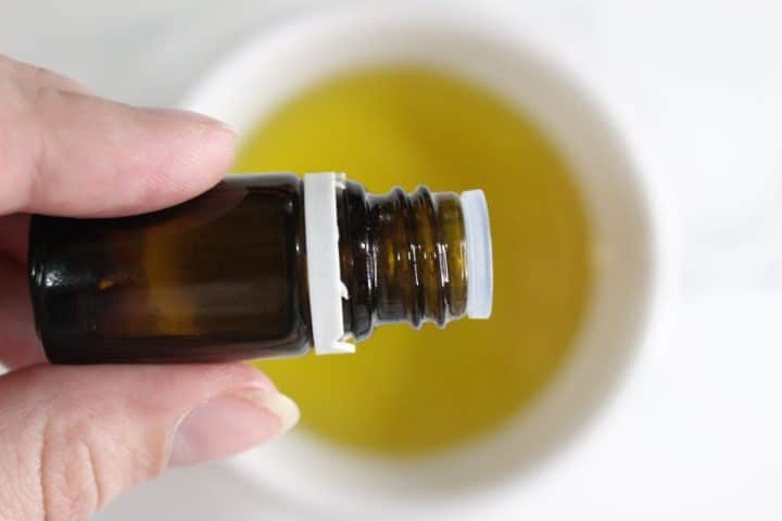 A hand holds an amber glass bottle with a white cap, positioned above a bowl containing yellow liquid.