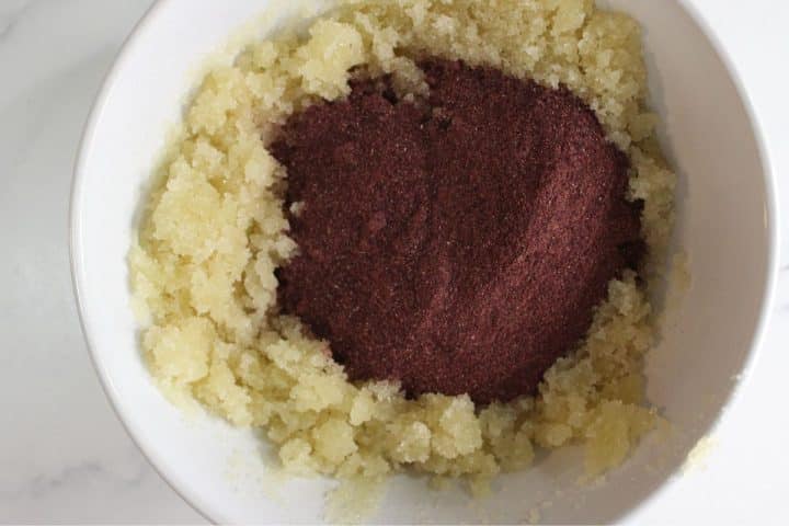 A white bowl containing a mixture of granulated sugar and a mound of dark purple powder, perfect for making your own DIY sugar scrub.