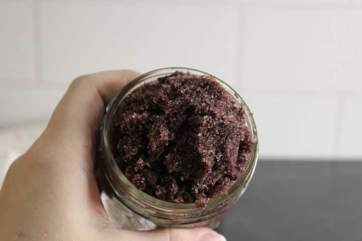 A hand holding a glass jar filled with a coarse, dark brown scrub mixture, likely a homemade sugar scrub, against a tiled background.