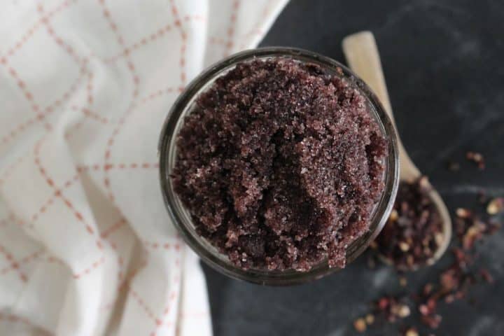 A jar filled with dark purple, coarse scrub sits on a black surface next to a wooden spoon and a white cloth with red lines.