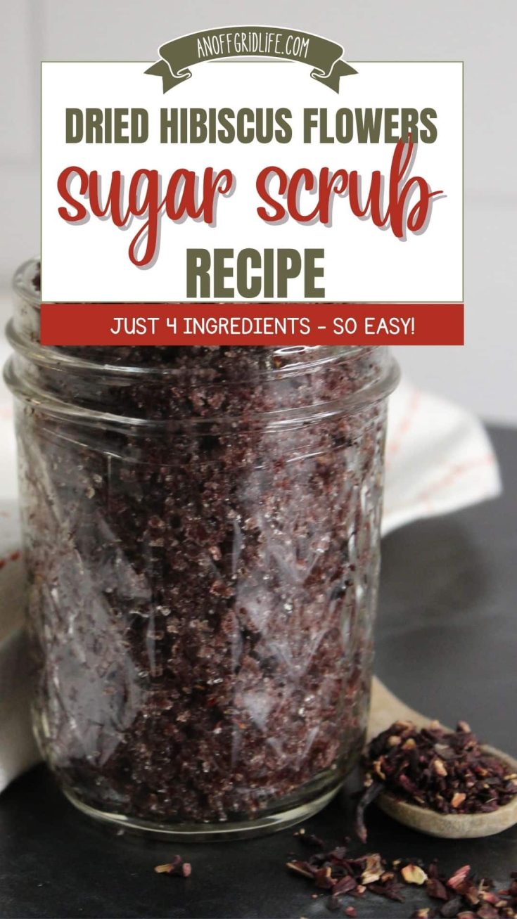 A glass jar filled with homemade dried hibiscus flower sugar scrub sits on a dark surface with a wooden spoon and dried hibiscus petals nearby.