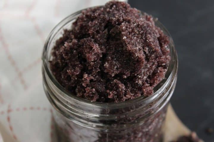 A glass jar filled with coarse, dark purple sugar scrub sits on a surface with part of a white cloth visible in the background.