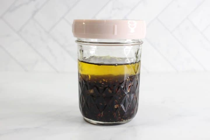 A glass jar with a light pink lid contains a dark liquid topped with a layer of yellow oil, set against a white tiled background.