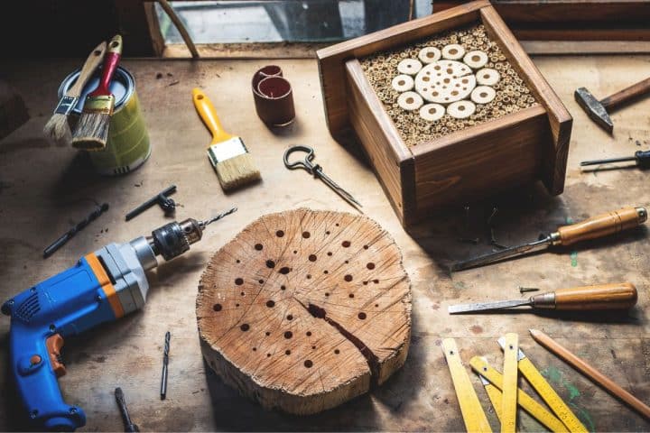 A woodworking bench with tools, a wooden block drilled with holes, a drill, paintbrushes, chisels, and a finished wooden insect hotel filled with bamboo tubes.